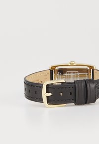 Montre en cuir avec un bracelet noir, boucle en métal doré et boîtier rectangulaire doré. Affichage clair avec des mécanismes internes visibles.