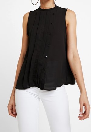 Blouse noire sans manches avec détails de boutons portée avec un pantalon blanc, montrant une partie du torse et des bras d'une personne.