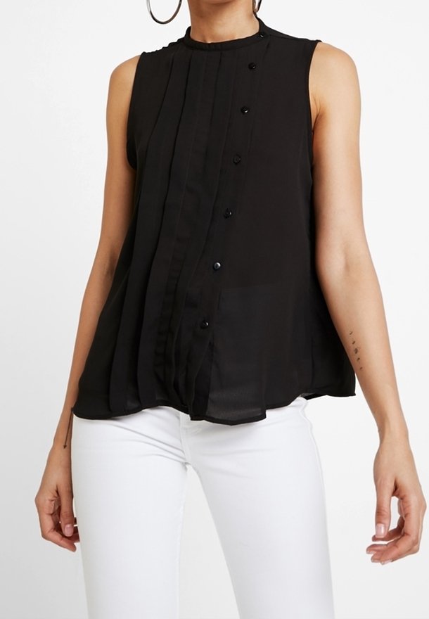 Blouse noire sans manches avec détails de boutons portée avec un pantalon blanc, montrant une partie du torse et des bras d'une personne.