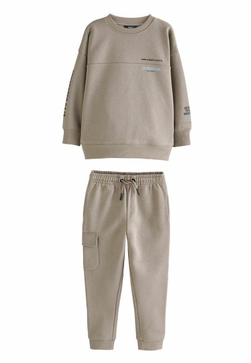 Beige fleece collegepaita ja siihen sopivat verkkarit, joissa on pyöreä kaula-aukko, pudotetut olkapäät, sivutaskut ja painettu teksti hihoissa.