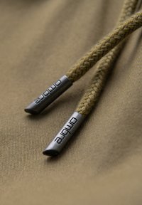 Gros plan d'un cordon tressé marron avec embouts métalliques gravés "ombre" sur fond de tissu kaki.