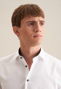 Camicia bianca con bottoni blu scuro, caratterizzata da una texture sottile e design con colletto aperto. Il modello ha capelli castani di lunghezza media.