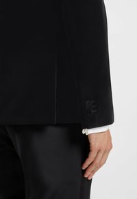 Zwarte fluwelen blazer met een gekartelde revers. Voorzien van drie knopen op de manchetten en een gladde, getailleerde pasvorm. Gstyled met een wit shirt.