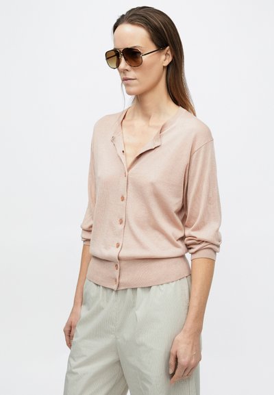 Frau trägt eine beige Strickjacke mit Knöpfen und hellgraue Hose, dazu braune Aviator-Sonnenbrille, steht vor einem einfarbigen weißen Hintergrund.