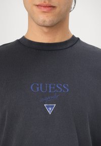 Jūras kara flotes īsais piedurkņu T-krekls ar izšūtu "GUESS" logotipu zilā krāsā, ar trīsstūra dizainu un vārdu "origins" uzrakstītu zem tā.