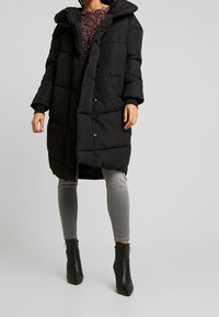 Schwarze Pufferjacke mit großem Kragen, Druckknöpfen und gesteppter Musterung. Getragen über einem burgunderfarbenen Strickoberteil und grauen, figurbetonten Jeans.