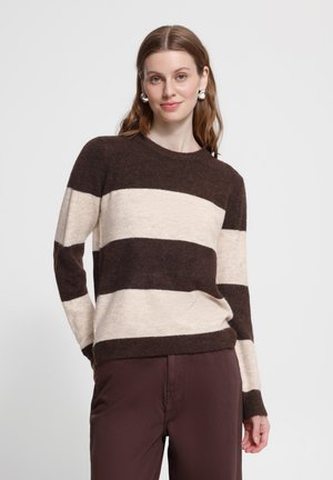 Donna dai lunghi capelli castani indossa orecchini d’argento vistosi, un maglione a righe orizzontali marrone e crema e pantaloni marroni, in piedi su uno sfondo semplice.