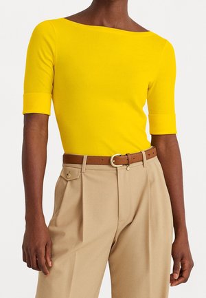 Strak zittend felgeel topje met korte mouwen, gecombineerd met beige hoge taillebroek en een bruine riem met een gouden gesp.