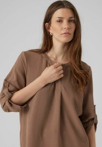 Blusa marrone, comoda e ampia, con maniche a tre quarti, polsini abbottonati, scollatura rotonda e pieghe sottili lungo il davanti.