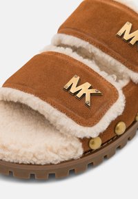 Sandalia de ante marrón con forro de borreguillo beige y logotipos dorados "MK" en las correas, con tachuelas de latón y suela de goma texturizada.