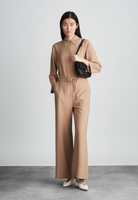 Svetlohnedá košeľa s dlhým rukávom a golierom, zladené široké nohavice a čierna crossbody taška. Štruktúrovaná tkanina a viazaný detail v páse.