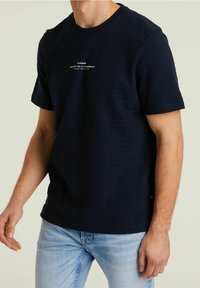 Marineblaue Kurzarm-T-Shirt mit strukturiertem Stoff, Rundhalsausschnitt und weißem Logodetail auf der Brust. Getragen mit hellblauen Jeans.