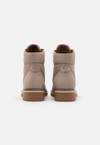 Botas de tobillo beige con un exterior de cuero suave, puntera redondeada y suela de madera marrón. Cuenta con un detalle de logo en el costado y un interior acolchado.