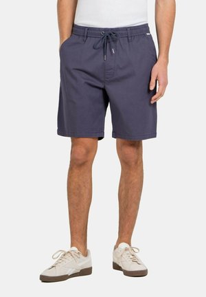 Hombre que lleva puestos pantalones cortos azul marino con cordón y zapatillas blancas, de pie sobre fondo blanco.