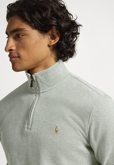 Young man with curly hair wearing a light green quarter-zip sweater with a small embroidered horse logo on the chest, facing left.