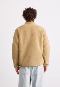 Beige Fleecejacke mit Kragen, strukturiertem Oberflächenmaterial und lässiger Passform, kombiniert mit hellblauen Jeans.