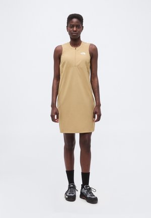 Persona de pie mirando hacia adelante, lleva un vestido sin mangas color beige hasta la rodilla, calcetines negros y zapatos de senderismo negros sobre un fondo claro y liso.