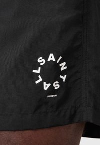 Mustat kangashousut, joissa on sileä pinta, ja joissa on ympyrän muotoinen valkoinen logo, jossa lukee "SAINT LONDON" hemman reunassa.