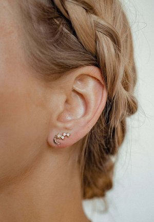Goldene Creolen zeigen ein Design in Form eines Halbmonds und drei kleine, klare Edelsteine, die in einem Cluster angeordnet sind, angesteckt an einem geflochtenen Haarhintergrund.