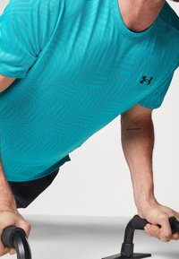 Turkos atletisk skjorta med geometrisk mönster, svart logga, bärs under armhävningar på handtag. Kort ärm, lättviktsmaterial.