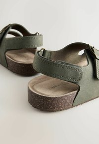 Grønne sandaler i faux læder med justerbare stropper, tekstureret overflade, korkfodbed og flad gummisål. Åbent design på siderne.