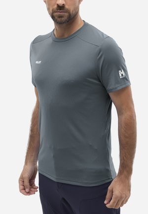 Homme portant un t-shirt de sport gris à manches courtes de marque Millet et un pantalon foncé, debout les bras détendus le long du corps.