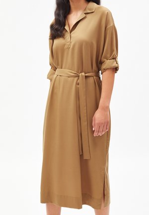 Kobieta w sukience do kolan w kolorze camel, z podwiniętymi rękawami, kołnierzykiem, paskiem wiązanym w talii oraz bocznymi rozcięciami na białym tle.
