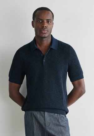 Mężczyzna w granatowym, krótkim, dzianym polo i szarych spodniach, stojący na tle jednolitej jasnej ściany z rękami za plecami.