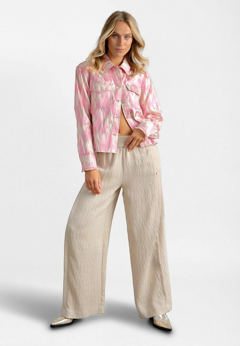 Femme posant en pantalon beige ample, chaussures pointues métalliques, et veste courte rose à motifs avec poches avant, sur fond blanc.