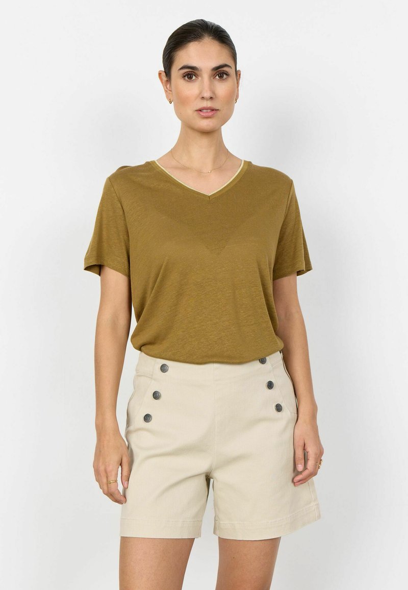 Femme portant un haut jaune moutarde à manches courtes et col en V ainsi qu'un short beige taille haute avec six boutons foncés, debout devant un fond blanc.