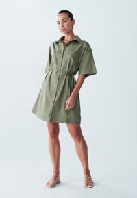 Oliivroheline särgimekko lühikeste varrukatega, nööbiga esiküljel ja vöökohtadega. Omab rinnaoidikuid ja vabalt langevat lõiget. Kanda minimalistlike sandaalidega.