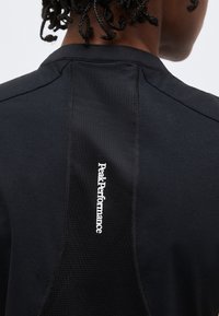 Οπίσθια όψη ενός ατόμου που φοράει μαύρο πουκάμισο με την επιγραφή «Peak Performance» τυπωμένη κάθετα στο πίσω κεντρικό πάνελ.