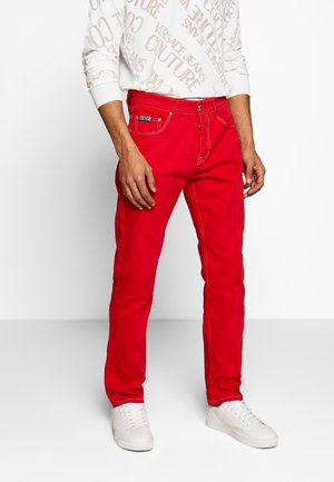 Mand iført lyse røde slim-fit jeans med hvide sømme, en hvid langærmet skjorte med grå tekst og hvide sneakers.