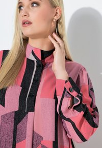 Langærmet bluse med et geometrisk mønster i pink, sort og grå. Fremstillet af glat stof med høj krave og sorte knapper.
