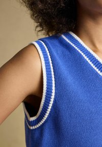 Varrukateta sinine kootud vest valge triibulise äärisega, mida kannab lokkide ja tumedate juustega inimene, nähtav õlast kuni rinnani.