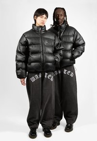 Black puffer jackets with a glossy finish, featuring a high collar and logo detail. Paired with loose dark gray pants and black footwear.