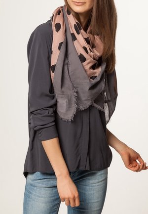 Femme portant une chemise gris foncé boutonnée, un jean bleu clair et une écharpe rose à pois noirs drapée autour du cou.