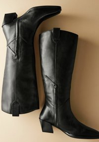 Botas de cuero negro hasta la rodilla con punta afilada y tacón apilado. Presentan detalles de cremallera y lengüetas traseras para facilitar su uso. Textura suave.