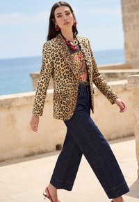 Femme marchant en plein air près de la mer, portant un blazer à imprimé léopard, un t-shirt à motif papillon, un jean à jambes larges, des boucles d'oreilles rouges, un collier rouge épais et des talons rouges.