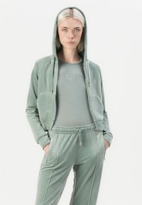 Groene velours trainingspak met een ritssluiting hoodie, een aansluitend topshirt met "Juicy" in rhinestones, en tapered broeken met een trekkoord in de taille.