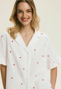 Biała koszula z krótkim rękawem i subtelnym paskowanym wzorem, ozdobiona rozrzuconymi czerwonymi motywami serc, z klasycznym kołnierzykiem.