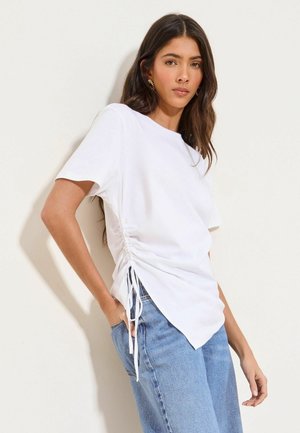 Pikkade tumedate juustega naine, kellel on kuldsed kõrvarõngad, valge külgedelt voltidega lühikeste varrukatega top ja sinised teksad, seisab tavalise heleda seina ees.