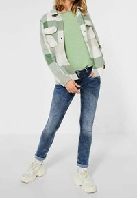 Zelená dlouhá rukávová košile pod bílou a zeleně kostkovanou košilí, spárovaná s modrými skinny džíny a robustními bílými teniskami.