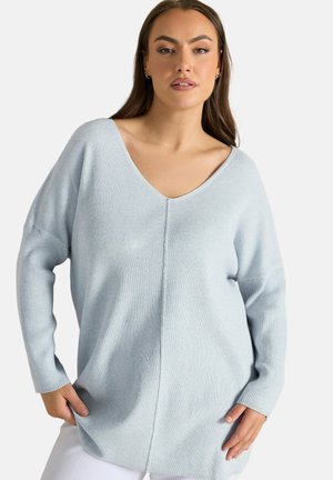 Kobieta ubrana w jasnoniebieski prążkowany sweter z dekoltem w serek i długimi rękawami, stojąca z rękami blisko kieszeni, na neutralnym tle.