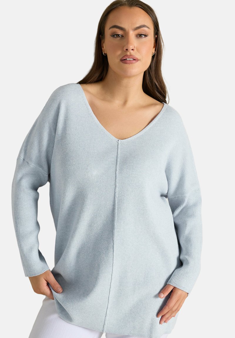 Donna che indossa un maglione a costine azzurro chiaro con scollo a V e maniche lunghe, in piedi con le mani vicino alle tasche, sfondo neutro.