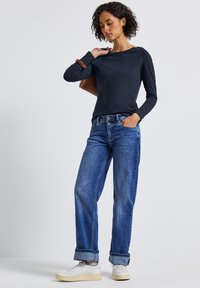 Frau mit kurzen, lockigen Haaren, die ein navyblaues Langarmshirt, umgeschlagene blaue Jeans, weiße Turnschuhe trägt und eine braune Handtasche hält.