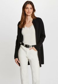Schwarze Strickjacke mit Reißverschluss, getragen über einem weißen Oberteil. Verfügt über lange Ärmel und eine glatte Textur, kombiniert mit eng anliegenden weißen Jeans und einem Gürtel.