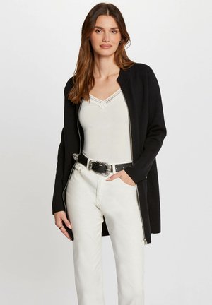 Cardigan noir avec un design zippé, porté sur un haut blanc. Présente des manches longues et une texture lisse, associé à un jeans blanc ajusté et une ceinture.