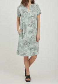 Lyhythihainen mekko vaaleanvihreänä, mustalla kukkakuviolla, jossa on V-pääntie, poimutuksia ja sivutaskut. Pehmeä, kevyt kangas.