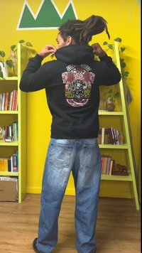 Black hoodie with colorful graphic design on the back, paired with light blue denim jeans, set against a yellow wall with shelves.